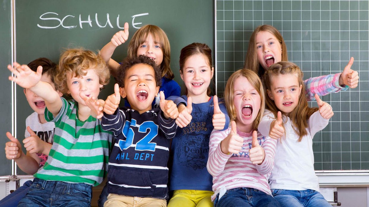 Kinder sitzen vor Tafel und halten Daumen nach oben Kinder sitzen vor Tafel und halten Daumen nach oben